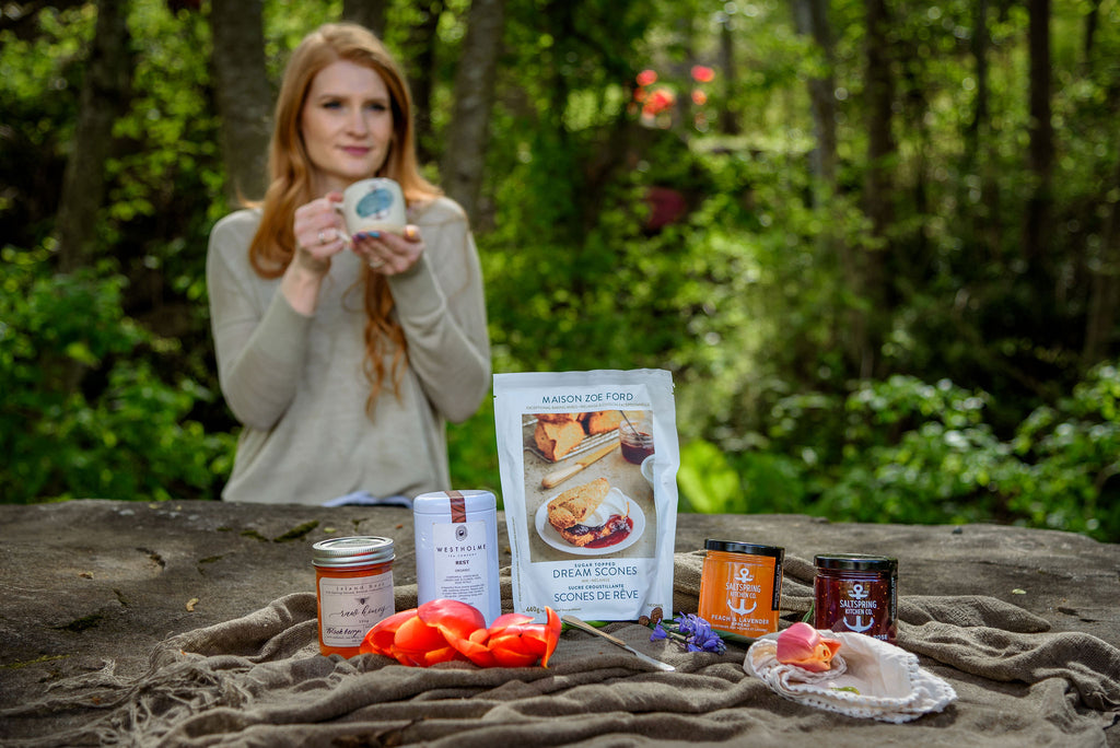 SaltSpring Kitchen Company Limited Edition Mother's Day Gift Boxes with jams, scone mix, honey and tea outside with woman drinking from a mug in the background