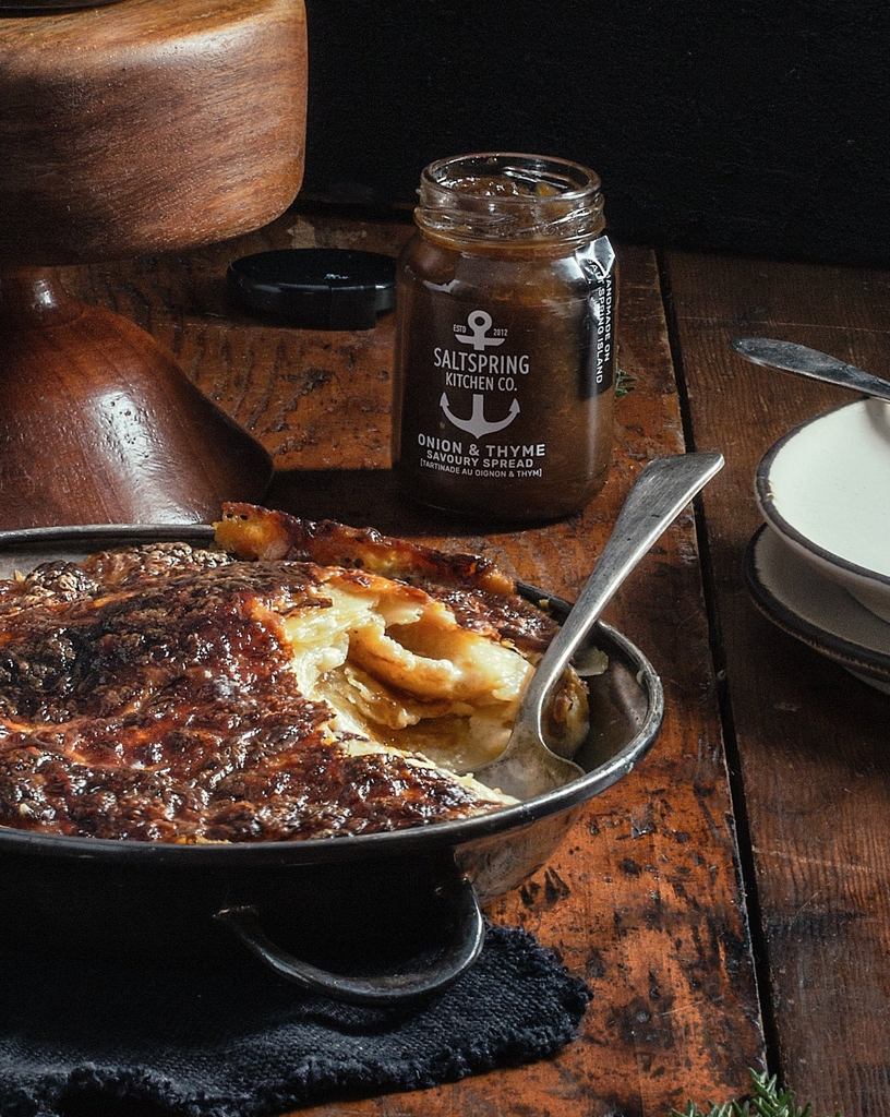 Baked dish of scalloped potatoes with a jar of SaltSpring Kitchen Co. Onion & Thyme Savoury Spread on a wooden table