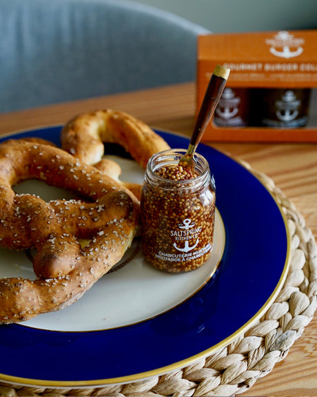 A small glass jar of SaltSpring Kitchen Co. Charcuterie Mustard sits on a blue and white plate with pretzels, on a woven placemat with a blurred kitchen background.