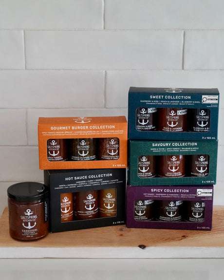 A collection of gourmet preserves from SaltSpring Kitchen Co. displayed on a wooden board against a white subway tile background. The products include Gourmet Burger Collection, Savoury Collection, Sweet Collection, Hot Sauce Collection, Spicy Collection, and a single jar of Tandoori Chutney.