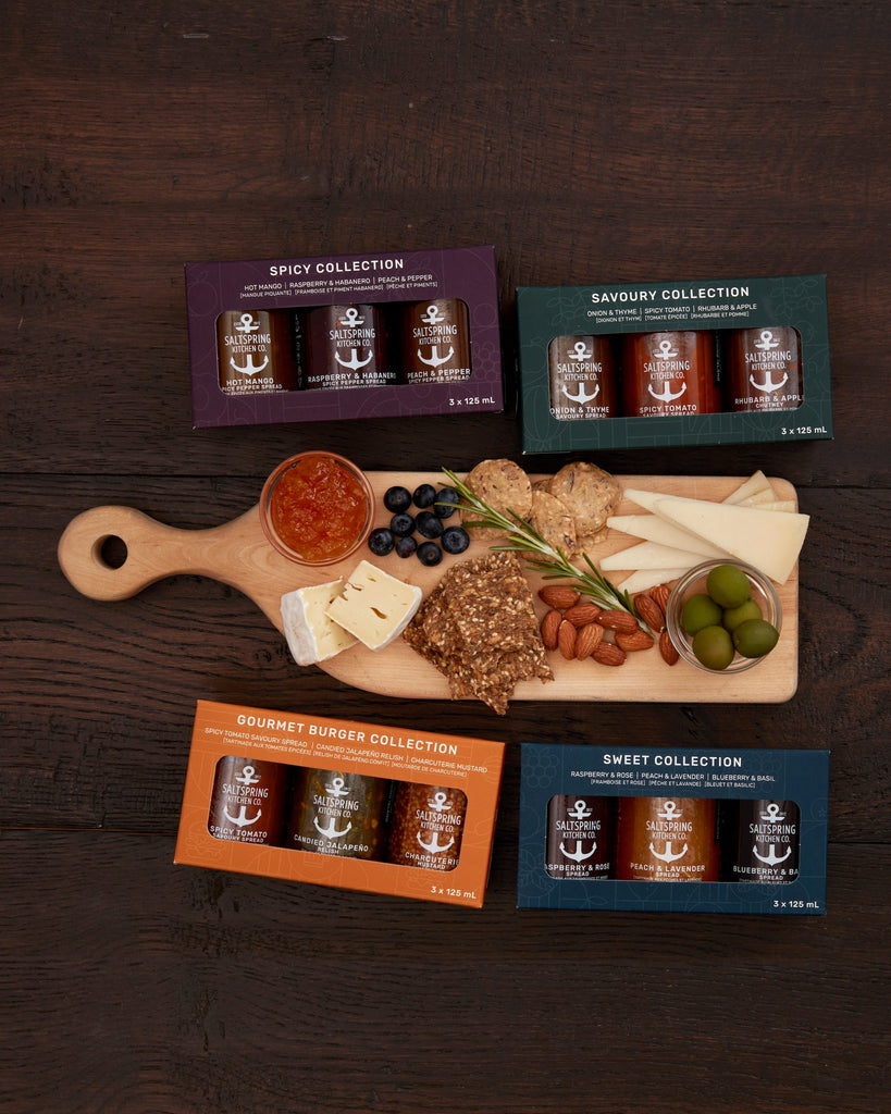 Collection of gourmet food items on a wooden board with packaging labeled 'Spicy Collection', 'Savory Collection', 'Gourmet Burger Collection', and 'Sweet Collection' on a dark wooden surface.