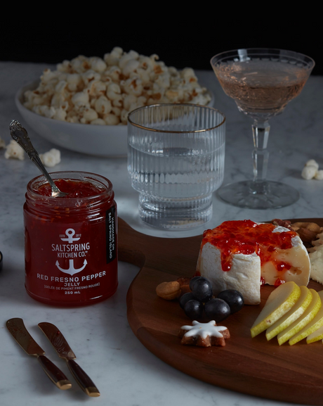 Plated appetizer with a jar of red Fresno pepper jelly, cheeseboard, and popcorn on a marble surface.