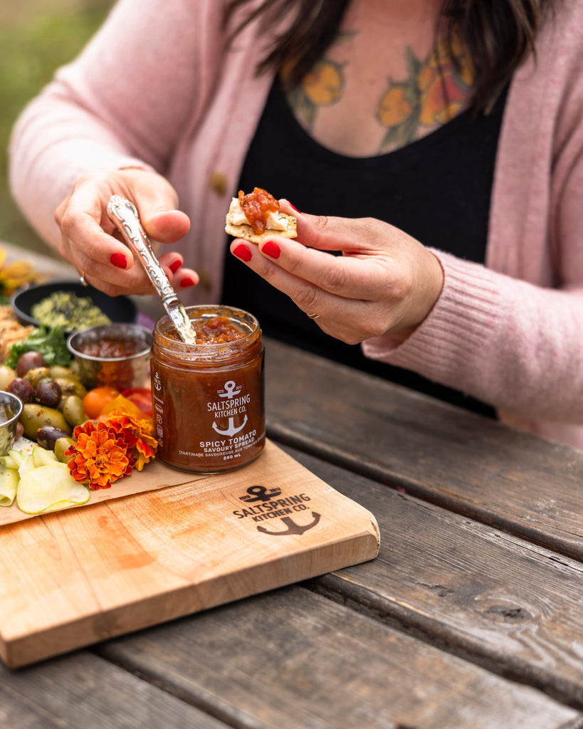 Person spreading a condiment on bread with a jar of it on a wooden board outdoors.