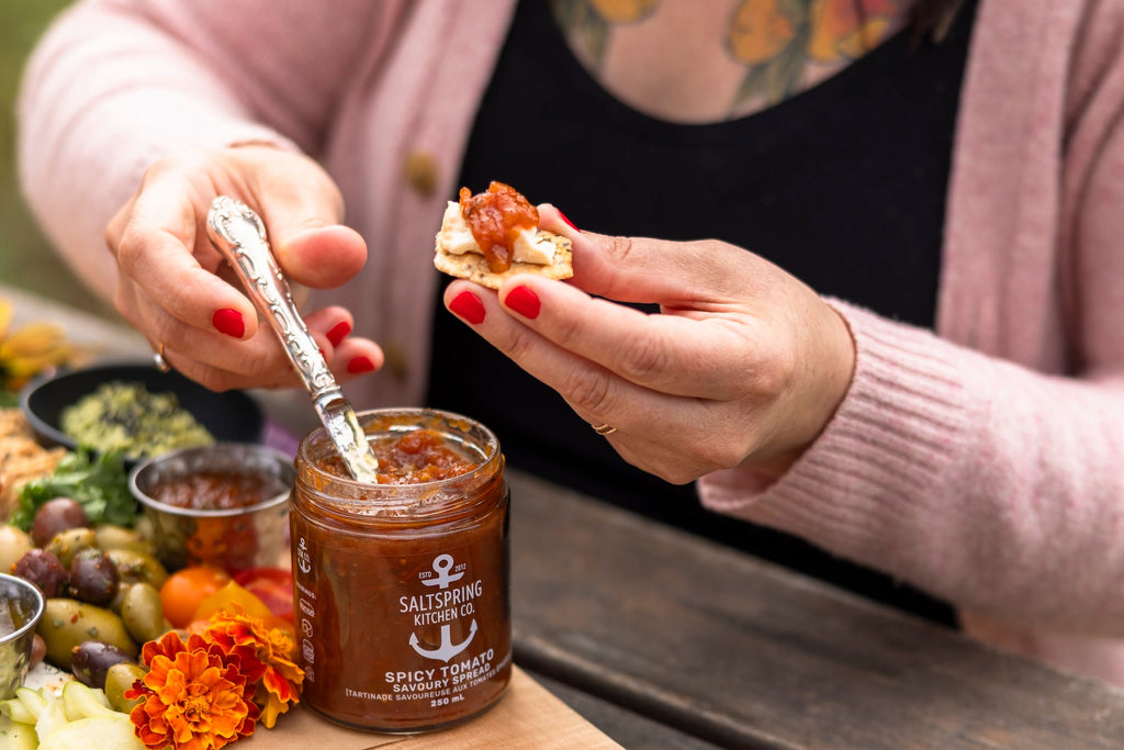 Person spreading a condiment on bread with a jar of it on a wooden board outdoors.